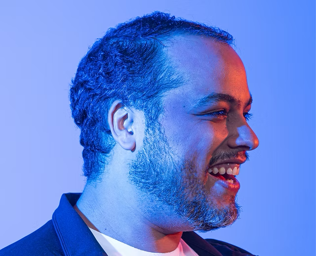 Retrato em perfil de Vinícius Meneses, autor de Pato no Galinheiro, com iluminação azul e magenta sobre fundo neutro.